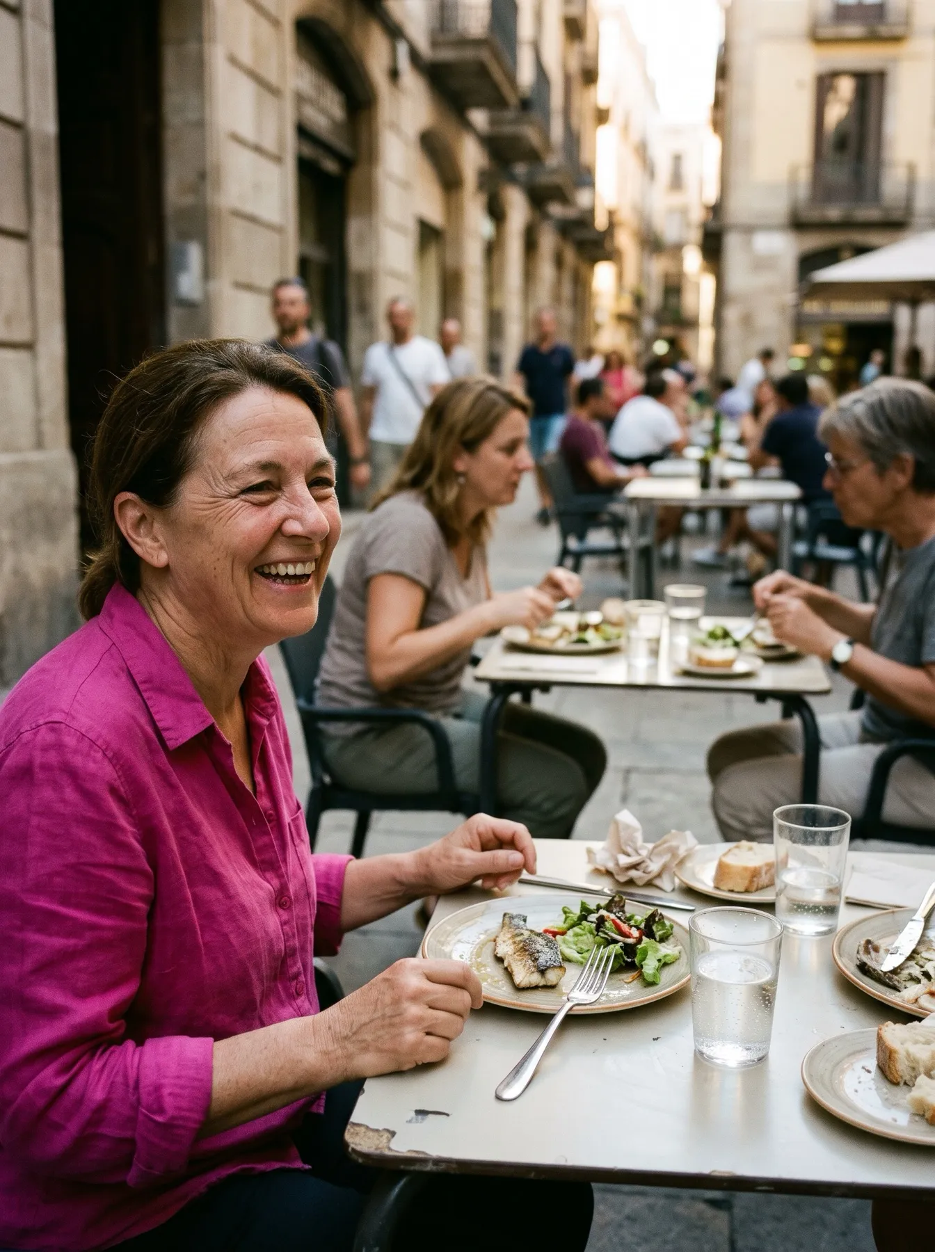 Una mujer con camisa rosa se ríe en una terraza con su plato de pescado y ensalada casi sin tocar y un vaso de agua delante